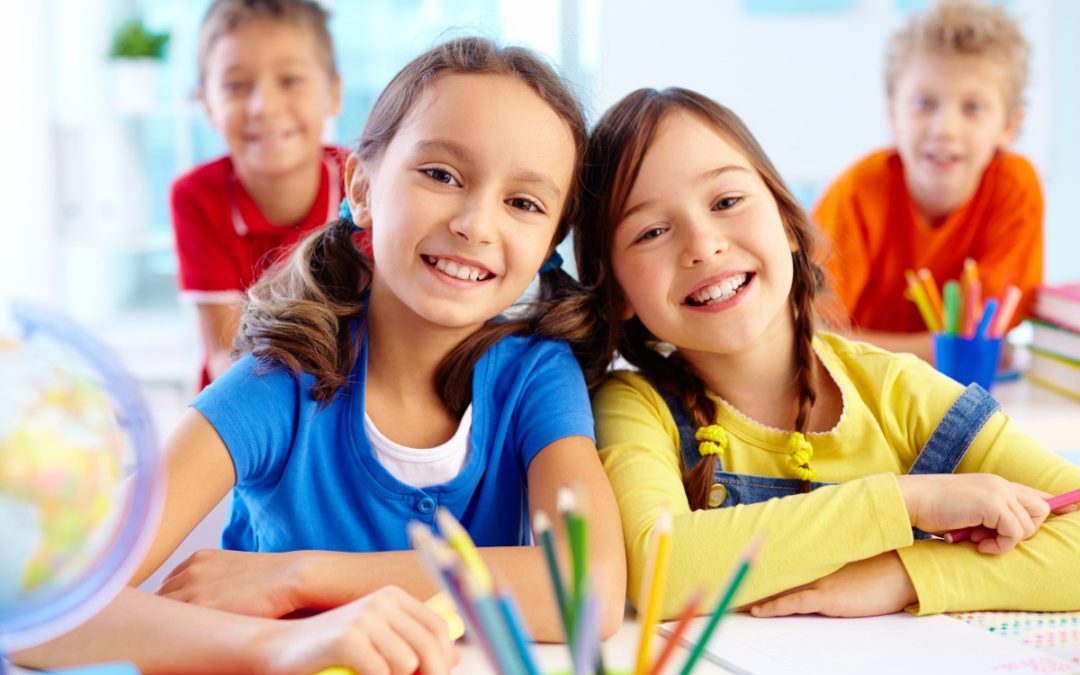 Four children smiling while drawing with colorful markers at a table in a classroom.